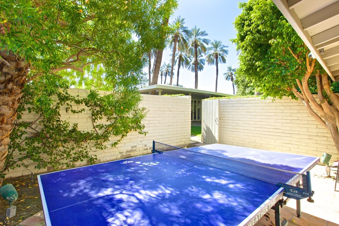 Blue table-tennis table on a walled patio shaded by trees, with palms beyond an open gate