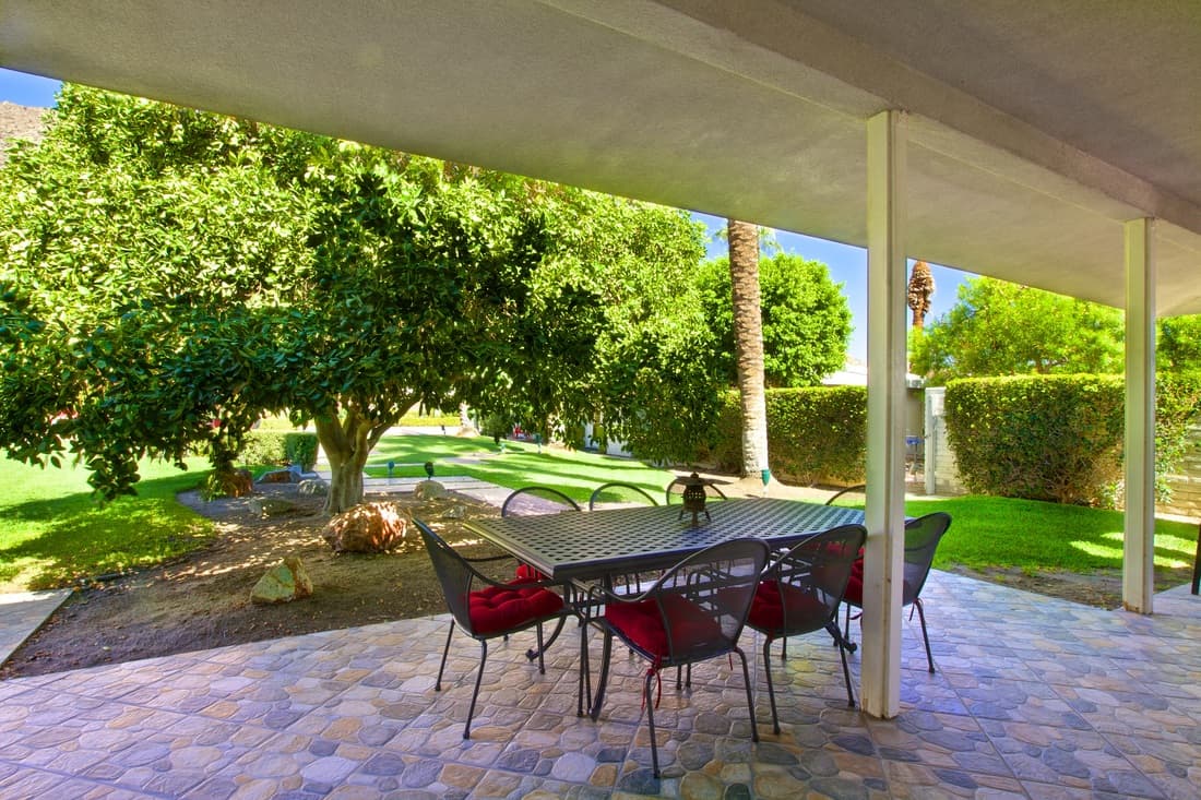 Covered patio with a black mesh table and red-cushioned chairs, looking out to a broad shade tree, clipped hedges and lawn
