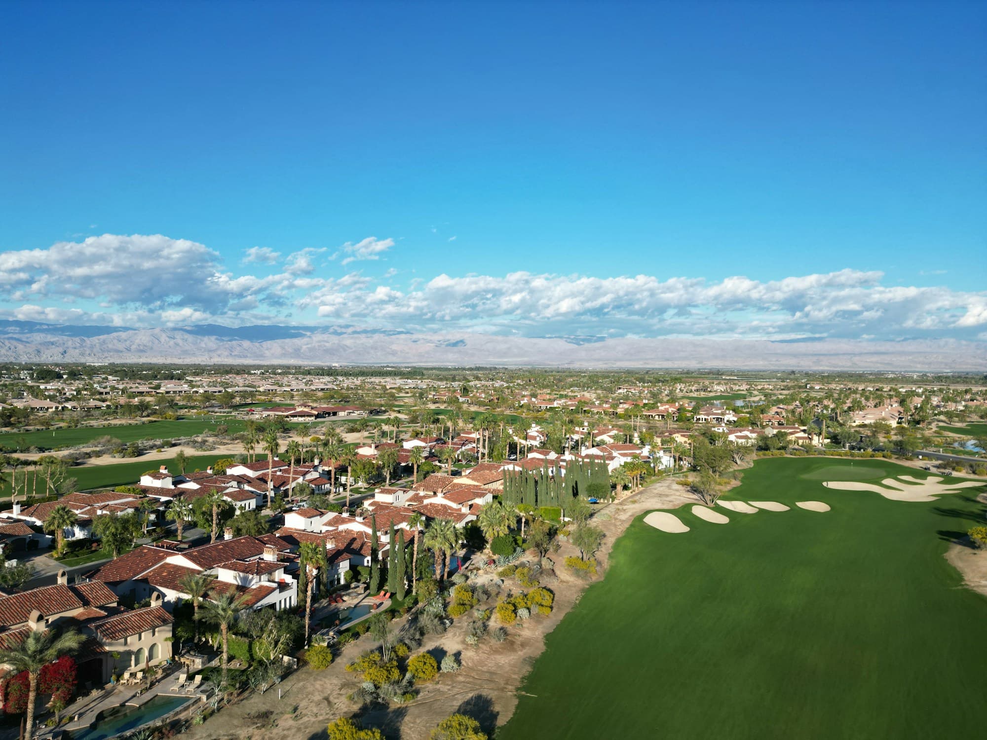 Aerial over a desert golf course: a green fairway and bunkers beside red-tiled homes, with mountains on the horizon