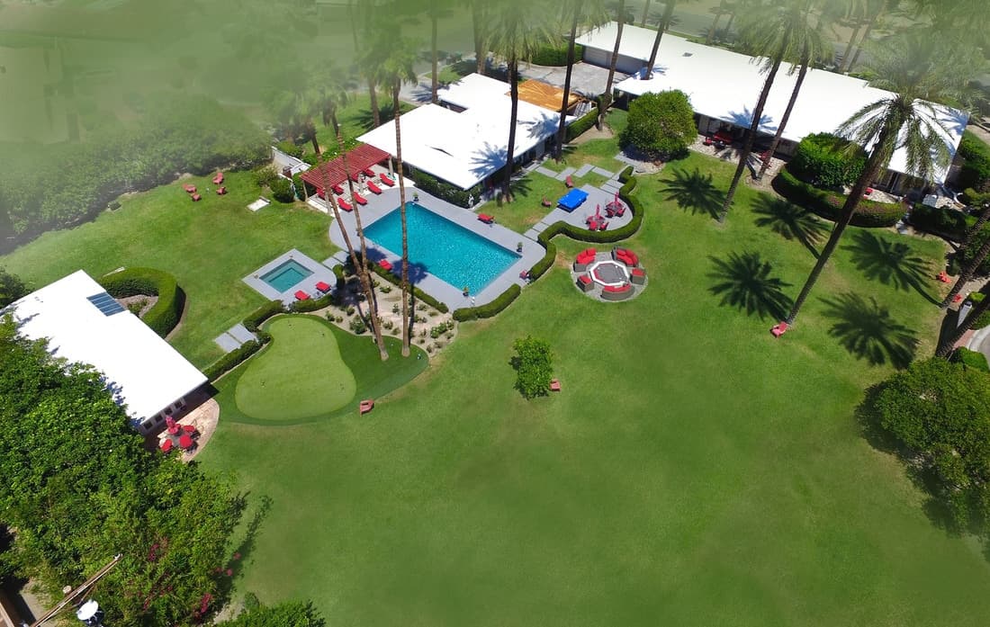 Aerial over the pool, spa, putting green and a red-seated fire pit, among date palms and three white-roofed buildings