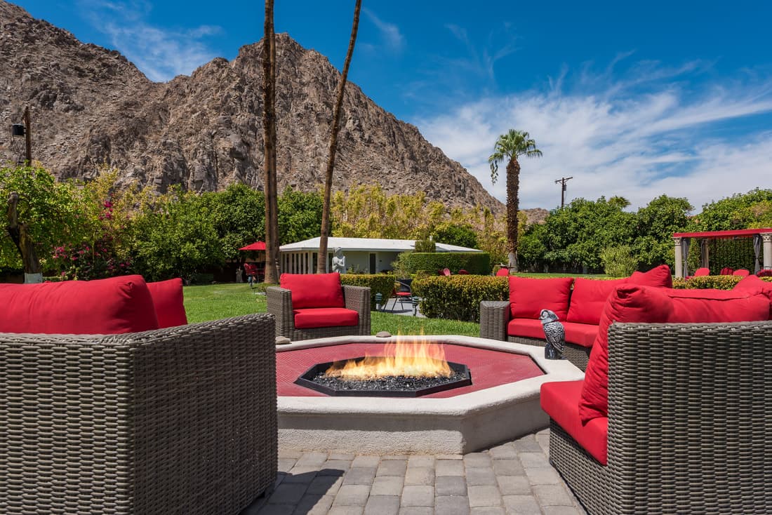 Burning octagonal fire pit ringed by red-cushioned wicker sofas, with a white cottage and a rocky mountain behind