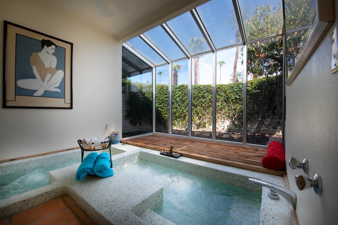 Sunken soaking tub in a glass-walled sunroom, with blue towels on the ledge and a clipped hedge outside