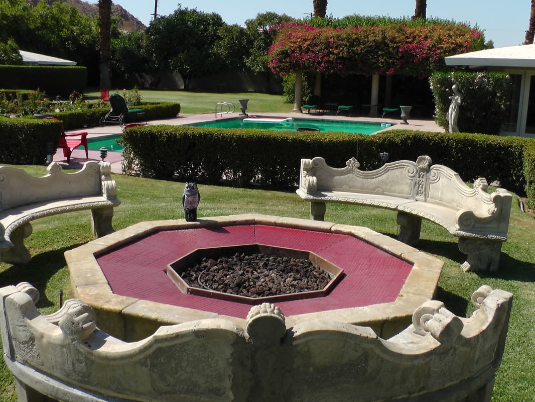 Octagonal fire pit with a red-tiled rim and a bed of dark stones, ringed by carved stone benches, with a pool beyond