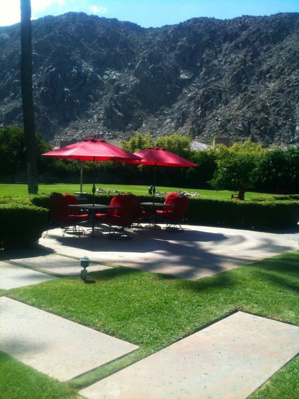 Two red umbrellas over patio dining sets beside a clipped hedge and lawn, with a rocky mountain behind
