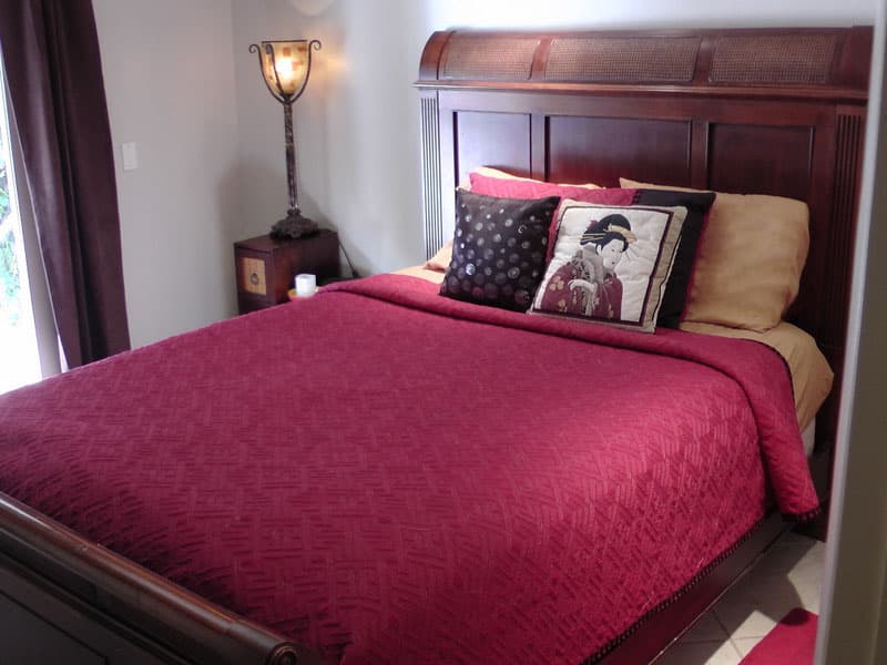 Bed with a quilted red cover and a tall carved wood headboard, beside a curtained window and a lit floor lamp
