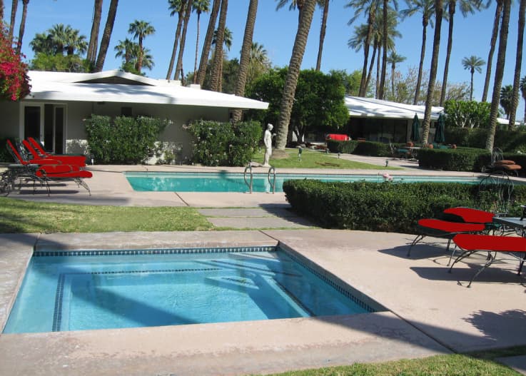 Square spa in the foreground, with a pool, red loungers, a stone statue and low white houses among palms behind