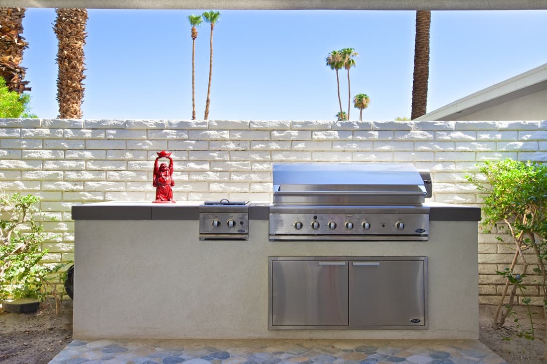 Built-in stainless barbecue grill and side burner in a pale counter, against a white block wall with palms beyond