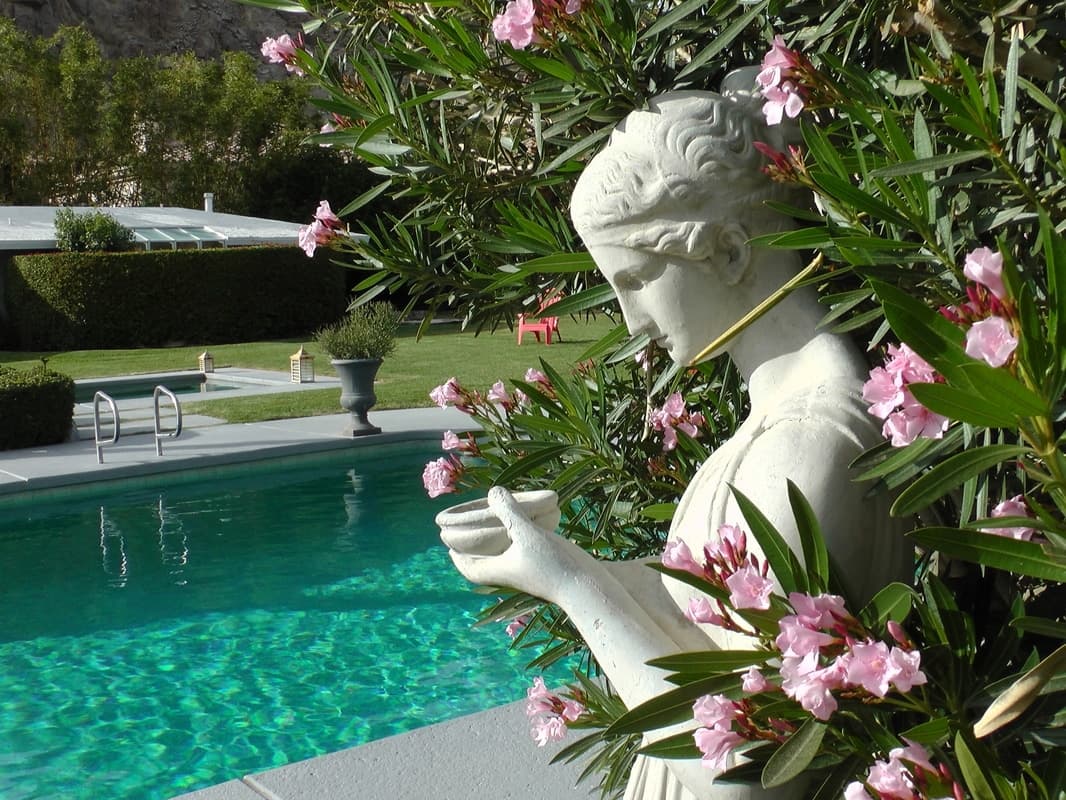 Carved stone statue of a woman holding a bowl among pink oleander, beside a turquoise pool and lawn