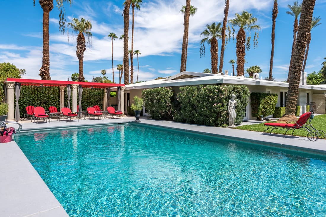 Rectangular pool beside a red-canopied pergola with red loungers, a stone statue and a low white house among palms