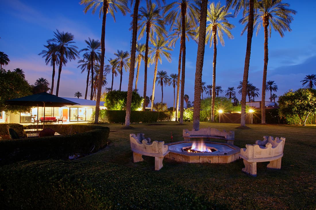 Burning octagonal stone fire pit and carved stone benches on a lawn, with date palms and a lit glass-walled house at twilight