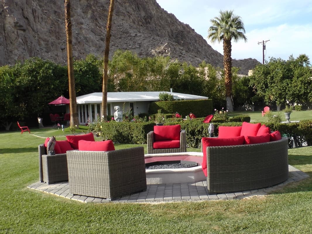 Curved wicker sofas with red cushions ringing an octagonal fire pit, with hedges, a low white house and a rocky slope behind