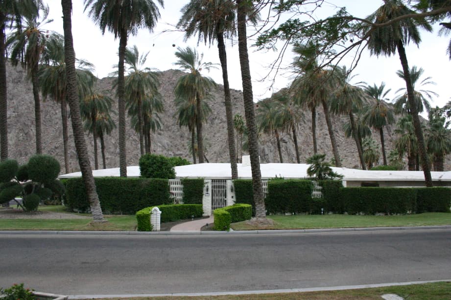Low white-roofed house behind a clipped hedge and gate, seen from the road through date palms, with a rocky mountain behind